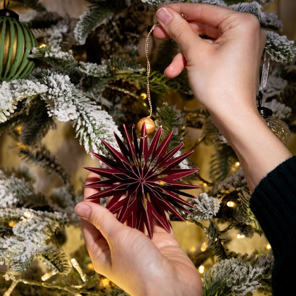 Hände halten einen sternförmigen Weihnachtsanhänger aus Papier vor einem künstlichen, verschneiten Weihnachtsbaum mit Beleuchtung.