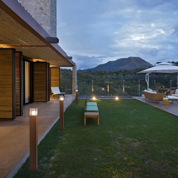Moderne Pollerleuchten in Bronze-Optik beleuchten einen Garten mit Rasen und Terrasse sowie Außenmöbeln vor einer Gebirgskulisse.