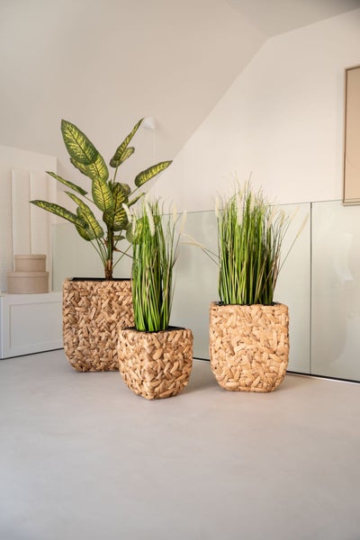 Künstliche Gräser und eine Blattpflanze in geflochtenen Übertöpfen in einem hellen, modernen Wohnraum mit Glasgeländer.
