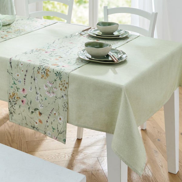 Gedeckter weißer Esstisch mit hellgrüner Tischdecke und floralem Tischläufer sowie grünem Geschirr in hellem Ambiente.