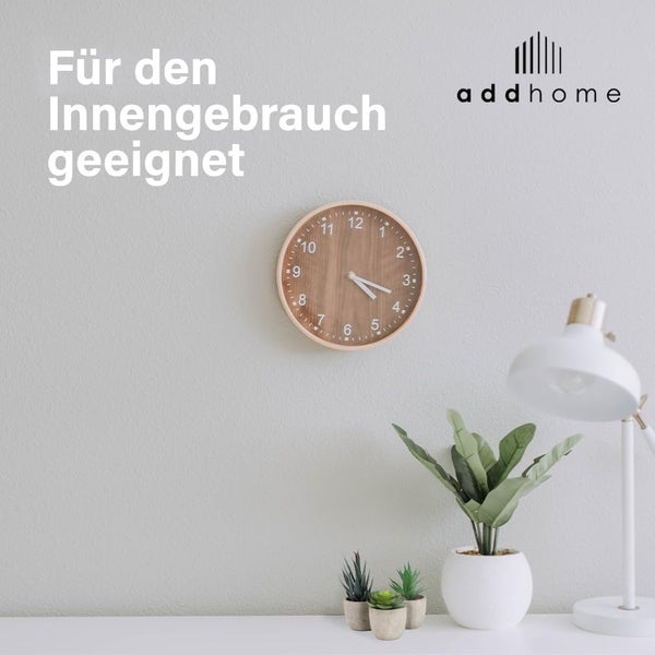 Runde Wanduhr in Holzoptik in einem hellen Innenraum neben einer Schreibtischlampe und Pflanzen. Geeignet für den Innengebrauch. addhome Logo.