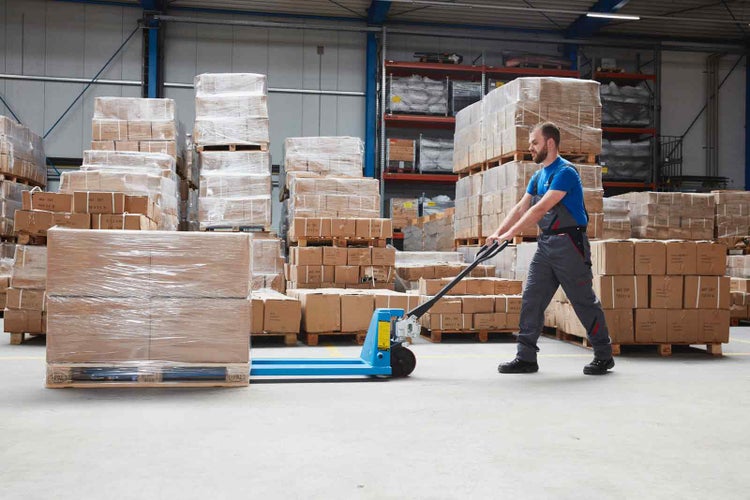 Ein Mitarbeiter in Arbeitskleidung zieht einen beladenen Hubwagen durch ein helles, organisiertes Warenlager.