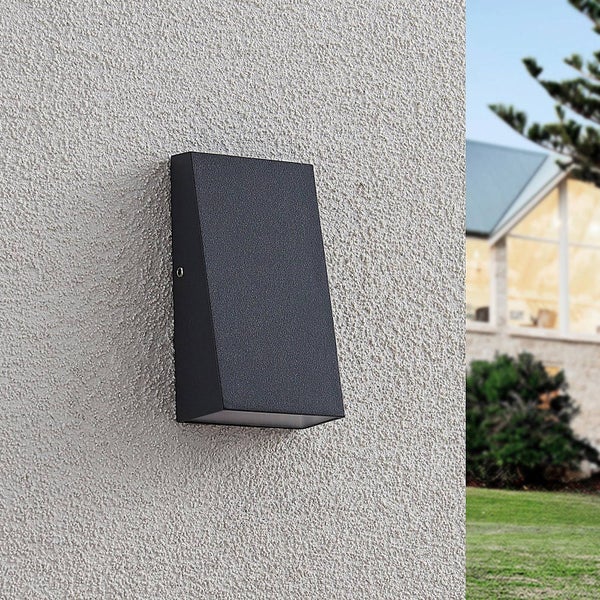 Moderne keilförmige Außenwandleuchte in Schwarz an einer strukturierten Hauswand im Außenbereich vor einem Garten.