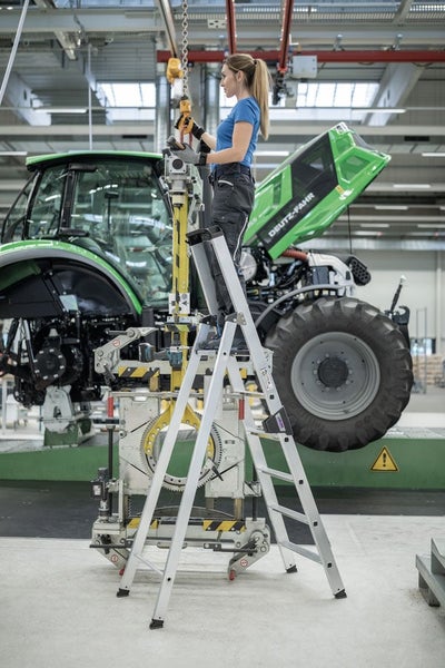 Eine Arbeiterin steht auf einer stabilen Aluminium Stufenleiter in einer Produktionshalle und bedient ein Hebegerät neben einem Traktor.