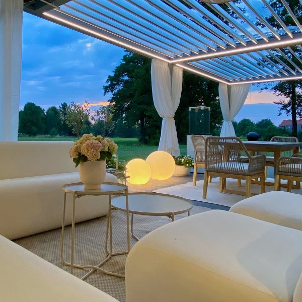 Modern gestaltete Terrasse mit beleuchteter Lamellenpergola, weißen Loungemöbeln, Esstisch und dekorativen Leuchtkugeln in der Dämmerung.