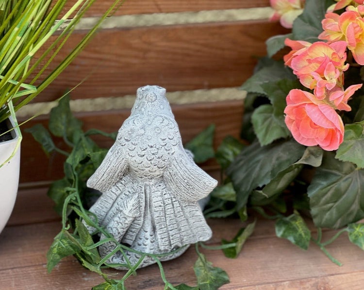 Gartenfigur Fisch aus Stein, platziert auf Holz zwischen grünen Blättern und rosa Blumen.