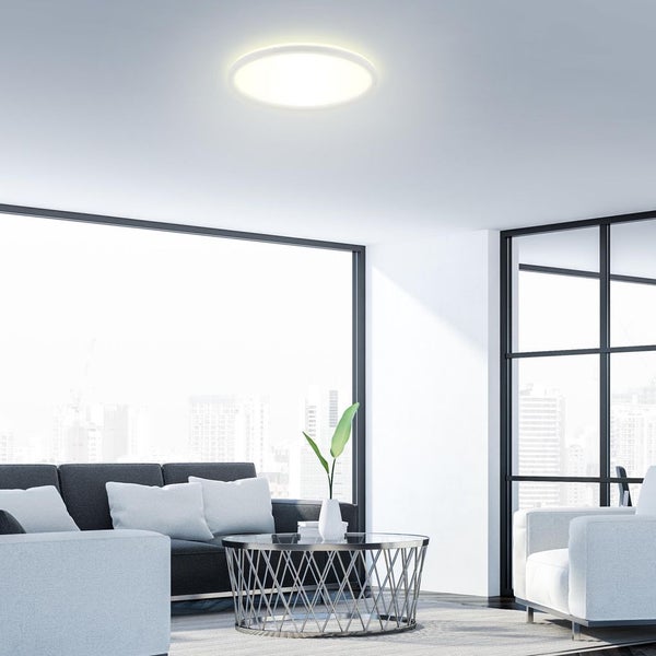 Moderne runde LED-Deckenleuchte in einem hellen Wohnzimmer mit Sofa und Couchtisch vor einem großen Fenster mit Stadtblick.