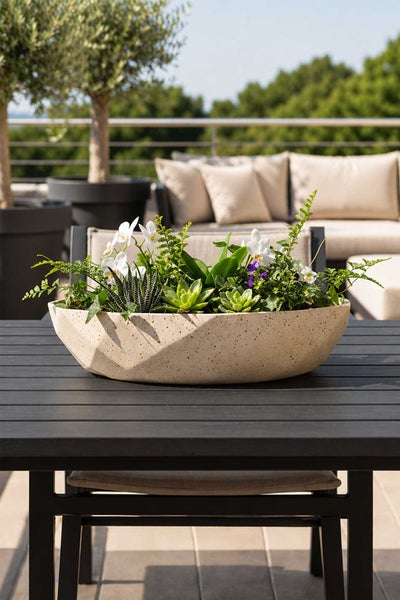 Moderne, bootförmige Pflanzschale mit gesprenkelter Textur auf einem Gartentisch, bepflanzt mit Sukkulenten und Blüten in hellem Gartenambiente.