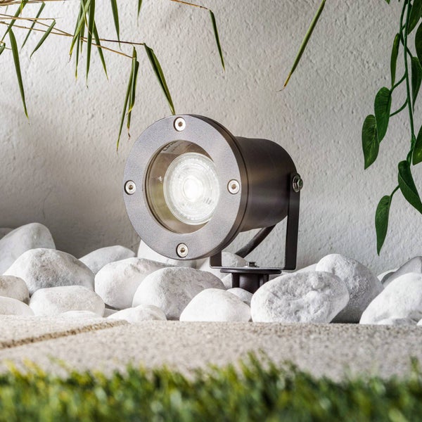 Schwenkbarer LED-Außenstrahler aus Metall, montiert in einem Beet aus weißen Steinen vor einer Hauswand mit Pflanzen.