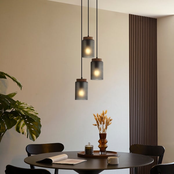 Dreiflammige Pendelleuchte mit zylindrischen Glasschirmen in unterschiedlichen Höhen über einem runden dunklen Esstisch in einem modernen Raum.