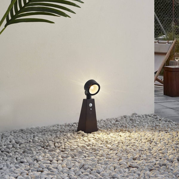 Moderner dunkelgrauer LED-Außenstrahler mit integriertem Bewegungsmelder, der hellgrauen Kies vor einer weißen Wand beleuchtet.