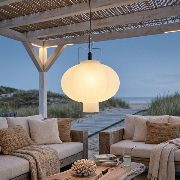 Ovale Pendelleuchte unter einer Holzpergola auf einer Strandterrasse mit Loungemöbeln aus Holz und Blick auf das Meer bei Dämmerung.