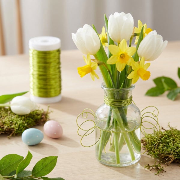 Frühlingshafte Tischdekoration mit Tulpen und Narzissen in einer Glasvase, geschmückt mit Dekodraht, Moos und bunten Ostereiern auf dem Tisch.