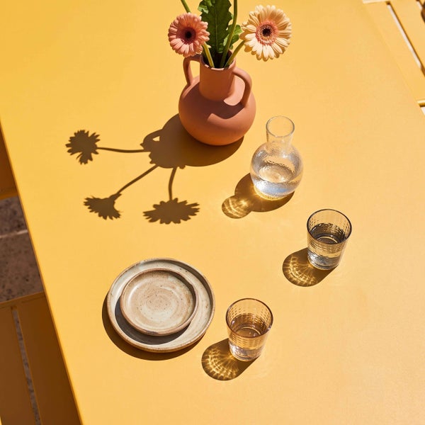 Gedeckter Gartentisch mit Keramiktellern, Glaskaraffe, Wassergläsern und einer Terrakottavase mit Blumen bei hellem Sonnenlicht.