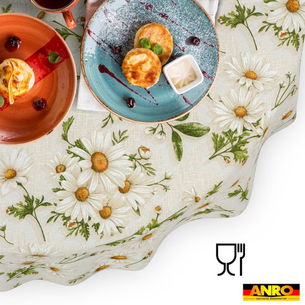 Tischdecke mit weißen Margeriten und grünen Blättern. Gezeigt mit Gedeck. ANRO Deutsche Manufaktur Logo und lebensmittelecht Symbol.