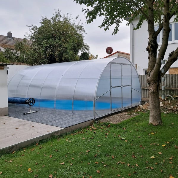 Überdachter Gartenpool mit einer halbrunden Schwimmbadüberdachung aus Kunststoff auf einer gefliesten Terrasse neben einer Rasenfläche.