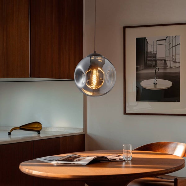 Moderne Pendelleuchte mit rauchgrauem Glasschirm über einem runden Holztisch in einer hellen Küche mit warmem Licht.