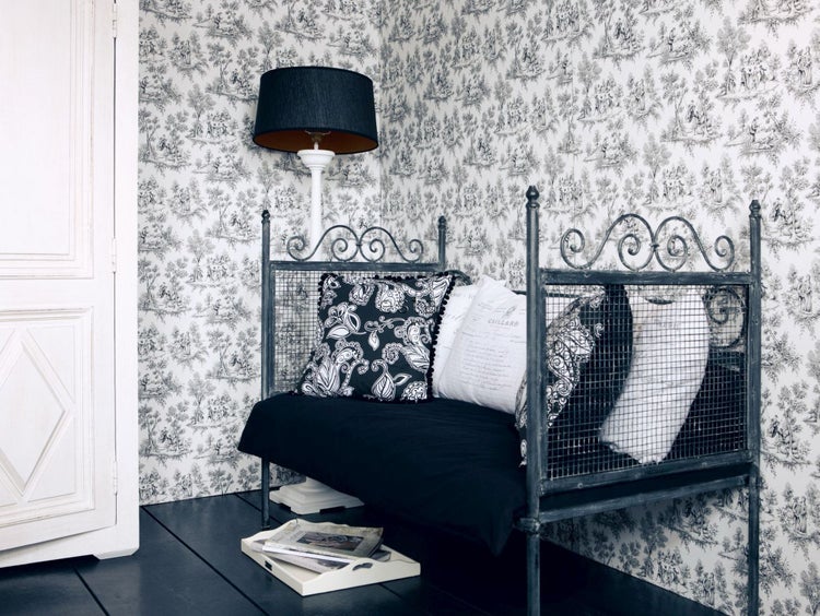 Tagesbett mit Metallgestell und Schnörkeln, gemusterten Kissen vor einer Wand mit Toile-de-Jouy-Tapete. Daneben steht eine Stehleuchte.