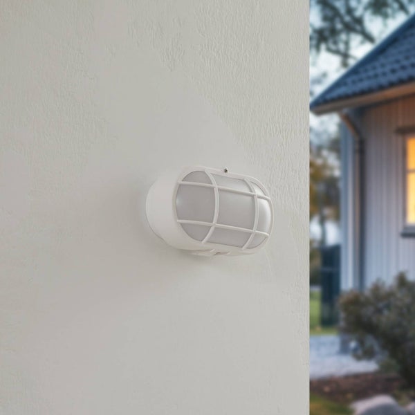 Ovale weiße Außenwandleuchte mit Schutzkorb und matter Abdeckung montiert an einer Fassade im hellen Außenbereich.
