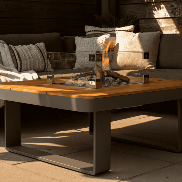 Quadratischer Feuertisch aus Holz und Metall mit Glasumrandung, Flammen und Steinen in einer gemütlichen Gartenlounge.