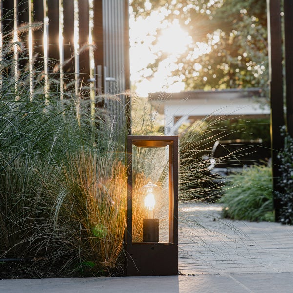 Moderne rechteckige Außenlaterne mit Filament-Leuchtmittel auf einem Steinweg neben Ziergräsern in einem sonnigen Garten.