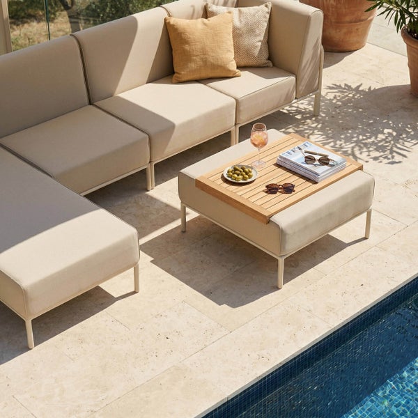 Helle Lounge-Ecke am Pool mit modularem Ecksofa, Polsterhocker mit Holztablett und Kissen auf einer Steinterrasse.