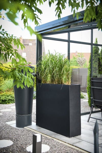 Moderner Garten mit einem hohen, rechteckigen Pflanzkübel in Anthrazit und Ziergras vor einem Wintergarten aus Glas.