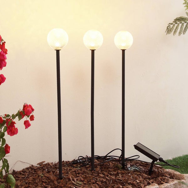 Drei leuchtende Solar-Gartenleuchten mit kugelförmigen Gläsern auf schwarzen Erdspießen in einem Beet mit Rindenmulch vor einer hellen Wand.