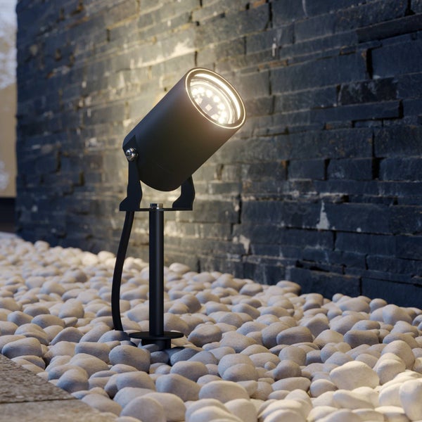 Schwarzer LED-Gartenstrahler auf einem Erdspieß in einem Beet aus weißen Kieselsteinen vor einer dunklen Steinwand.
