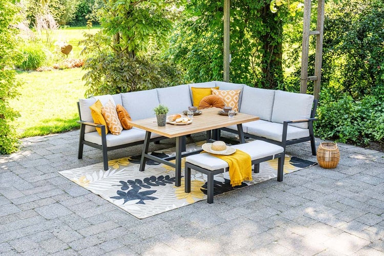 Garten-Lounge-Set auf Steinterrasse mit Ecksofa, Holztisch, Bank, grauen Polstern und Outdoorteppich in einem hellen, begrünten Garten.