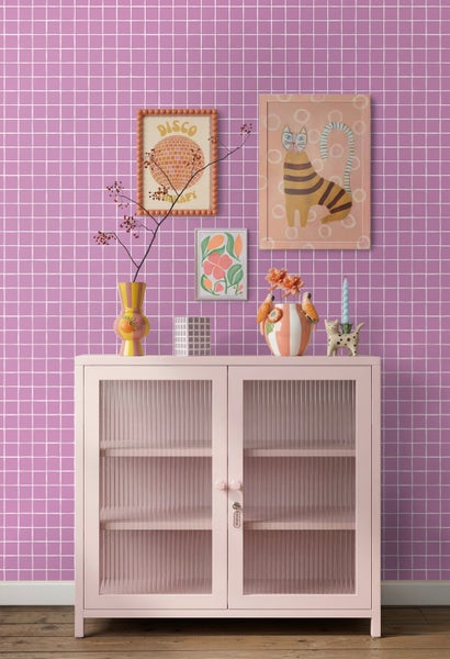 Rosa Metallschrank mit Riffelglastüren vor lila Tapete mit weißem Karomuster, dekoriert mit bunten Vasen und Wandbildern.
