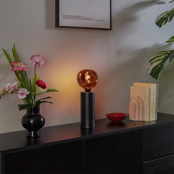 Moderne schwarze Tischleuchte mit dekorativer LED-Filament-Lampe auf einer dunklen Kommode neben Blumen und Büchern.