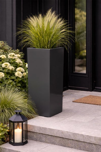 Hoher quadratischer Pflanzkübel in Anthrazit mit Ziergras auf einer Terrasse neben einer Laterne und Hortensien.
