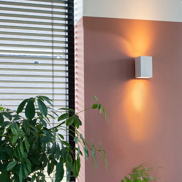 Quadratische Wandleuchte neben Jalousien und Zimmerpflanze