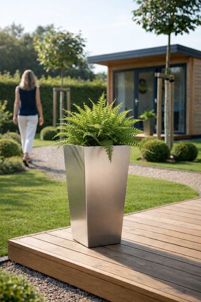 Hoher, quadratischer Pflanzkübel aus Edelstahl mit einem Farn auf einer Holzterrasse in einem hellen Garten mit Gartenhaus.