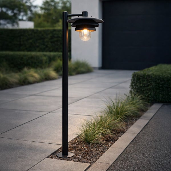 Schwarze Außenstehleuchte mit Schirm und leuchtender Glühbirne an einer modern gepflasterten Einfahrt mit Gras und Hecken.