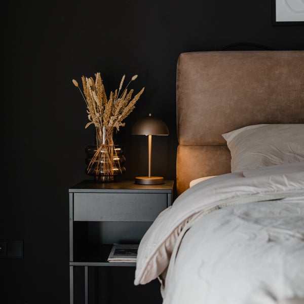 Schlafzimmer mit dunklem Nachttisch, pilzförmiger Metall-Tischleuchte, Glasvase mit Trockenblumen und braunem, gepolstertem Bettkopfteil.