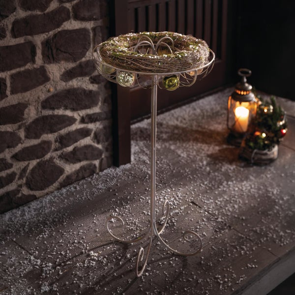 Weihnachtlicher Metallständer mit Zweigkranz und Weihnachtskugeln vor einer Steinwand mit einer Laterne als Weihnachtsdekoration.