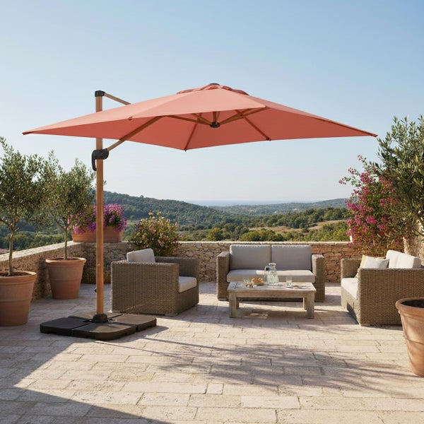 Großer Ampelschirm auf einer Steinterrasse mit Rattan-Sitzgruppe, Holztisch und Olivenbäumen in Kübeln vor einer Hügellandschaft.