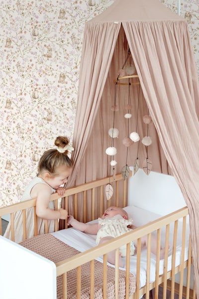 Kinderzimmer mit Baby im hölzernen Gitterbett unter einem Baldachin, davor ein Kleinkind, im Hintergrund Tapete mit Hasen- und Blumenmuster.