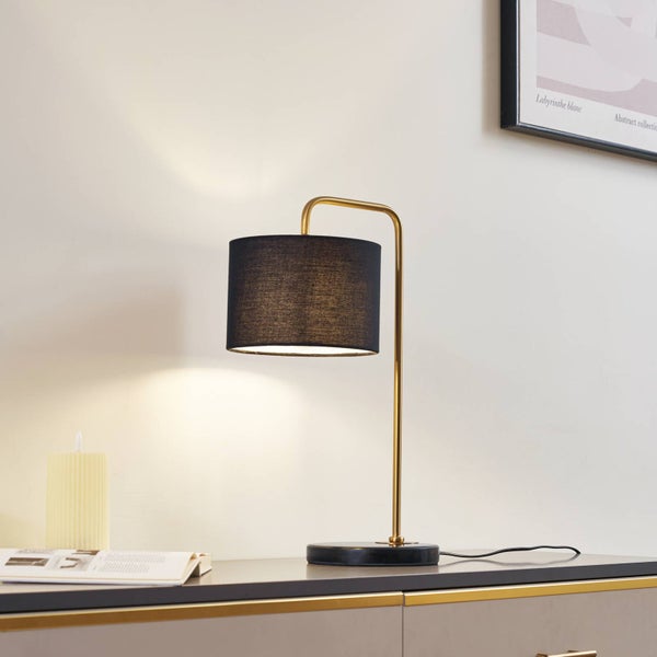 Moderne Tischleuchte mit schwarzem Stoffschirm, geschwungenem goldfarbenem Gestell und schwarzem Fuß auf einem Sideboard in hellem Raum.