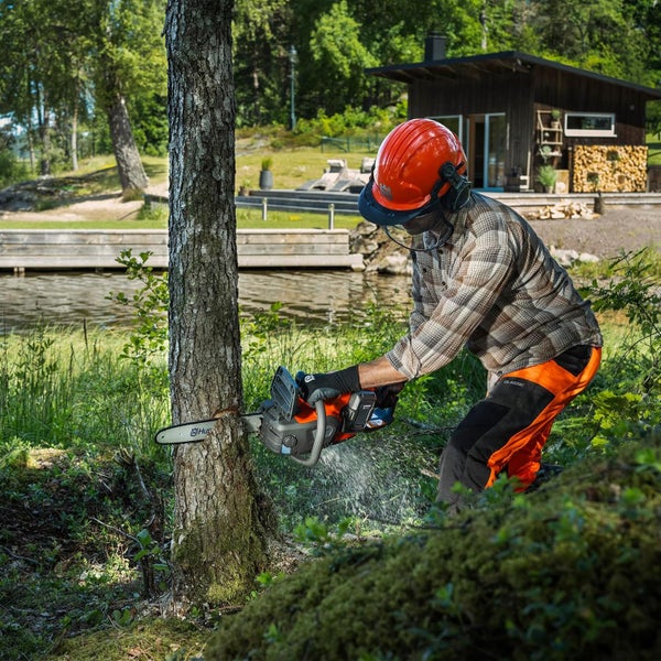 Person mit Schutzausrüstung sägt einen Baumstamm mit einer Husqvarna Akku Kettensäge in einer Waldumgebung am See.