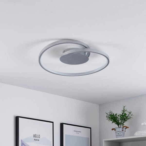 Moderne LED Deckenleuchte aus silberfarbenem Metall in Form von zwei ineinander verschlungenen Ringen an einer weißen Zimmerdecke.