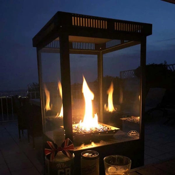 Muztag Gas-Feuerstelle mit schwarzem Metallrahmen und Glasscheiben, brennende Flamme auf blauen Glassteinen, auf einer Terrasse bei Nacht, Muztag Logo.