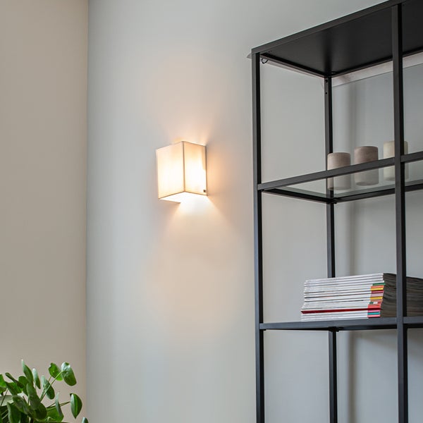 Moderne Wandleuchte in Würfelform, montiert an einer hellen Wand neben einem schwarzen Metallregal mit Zeitschriften.