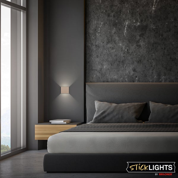 Moderne quadratische Wandleuchte mit Licht nach oben und unten in einem minimalistischen Schlafzimmer mit Polsterbett. Sticklights by Briloner Logo.