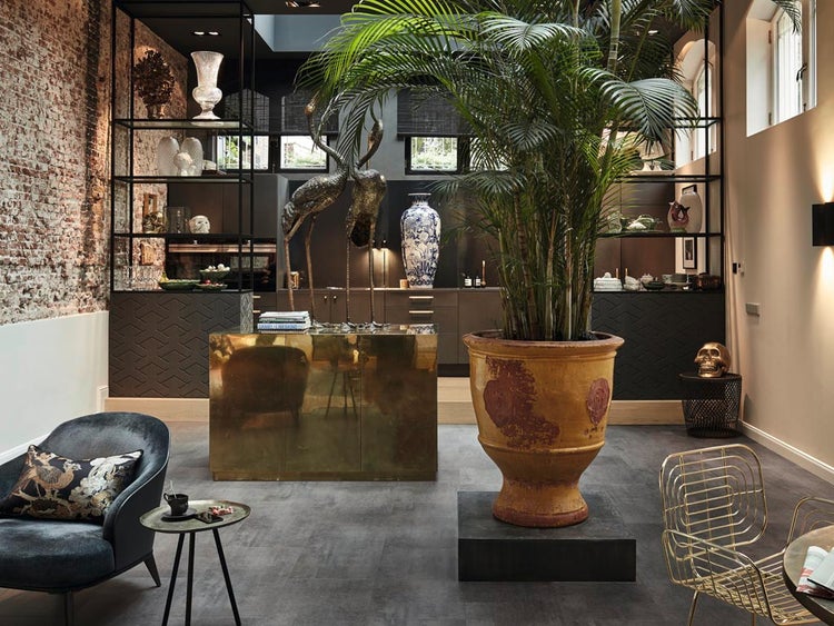 Exklusives Wohnzimmer mit moderner Einbauküche, großen Zimmerpflanzen, dekorativen Skulpturen und einer Wand mit freiliegendem Mauerwerk.