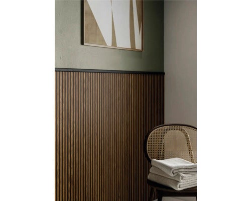 Moderner Innenraum mit Wandpaneelen aus dunklem Holz, grüner Wand, abstraktem Bild und einem Rattanstuhl mit gefalteten Handtüchern.