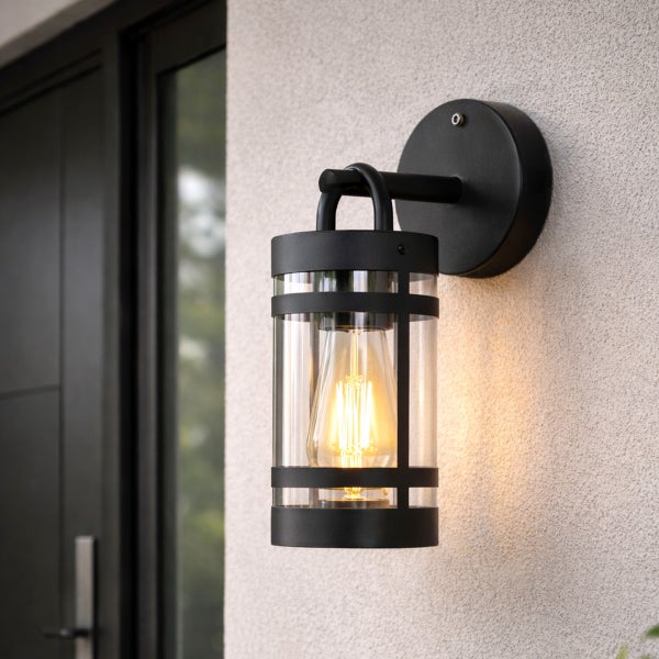 Schwarze Außenwandleuchte im Industriedesign mit klarem Glaseinsatz und leuchtendem Filament-Leuchtmittel an einer Außenwand montiert.
