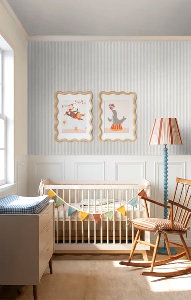 Kinderzimmer mit gestreifter Tapete, Wandpaneelen, Babybett mit Wimpelkette, Schaukelstuhl und Stehlampe.
